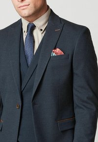 Granatowy garnitur z teksturowanej tkaniny, z klapami w kształcie notched, brązowym guzikiem oraz chusteczką w kieszeni w czerwone i turkusowe akcenty.