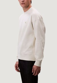 Světlý béžový svetr s kulatým výstřihem a žebrovanými manžetami, s malým zlatým logem na hrudi. Hladká látka s volným střihem.