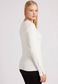 Maglione bianco a maniche lunghe con design aderente, polsini a coste e sottili dettagli con bottoni ai polsi. Tessuto morbido e superficie liscia.