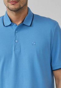 Helles blaues Polo-Shirt aus strukturiertem Stoff. Verfügt über einen klassischen Kragen mit dunkelblauer Einfassung und zwei Knöpfen sowie ein gesticktes Logo.