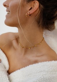 Goldene Creolen und ein zarter goldener Choker mit kleinen hängenden Charms, getragen mit einem softweißen Fleece-Wickel, der einen warmen Hautton zur Geltung bringt.