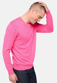 Homme en pull rose, jean foncé, souriant et touchant ses cheveux courts avec sa main droite, devant un fond uni.