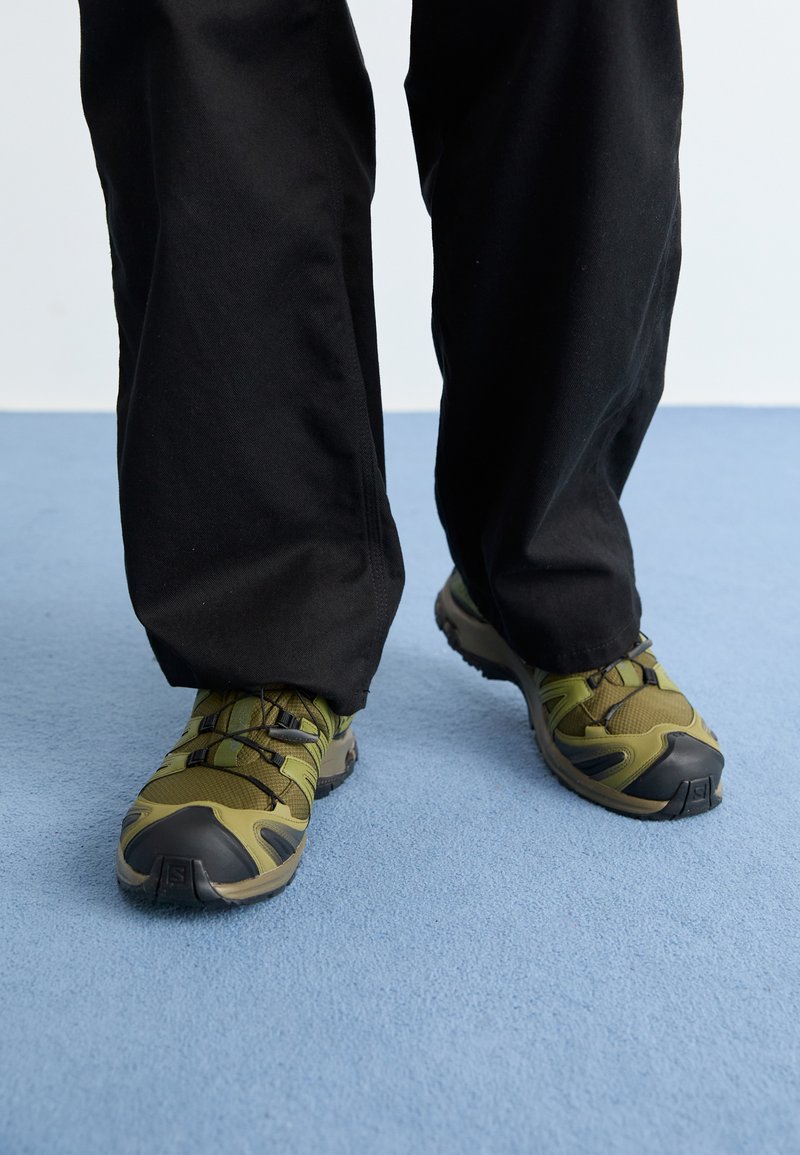 Πράσινα και γκρι αθλητικά παπούτσια με μαύρες λεπτομέρειες, με άνω μέρος από πλέγμα, λάστιχο σόλας και ελαστικά κορδόνια, φοριούν�ται με μαύρο παντελόνι πάνω σε μπλε χαλί.