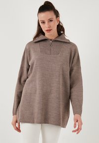 Brauner Strickpullover mit hohem Kragen und einem halben Reißverschluss, ausgestattet mit einem strukturierten Einsatz an der Brust und einem lässigen Schnitt. Lange Ärmel vorhanden.