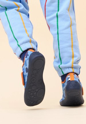 Blauwe kinderschoenen met een gestructureerde rubberen zool, voorzien van een patroon en een oranje bandje. G gedragen met lichtblauwe gestreepte broek.