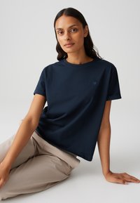 Maglietta in cotone blu navy con vestibilità comoda, scollo rotondo, maniche corte arrotolate e un sottile logo a forma di cuore sul petto.