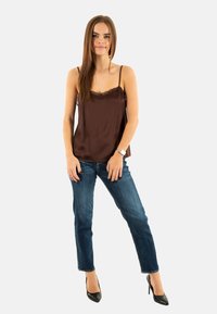 Camisola in satin marrone con bordi in pizzo, abbinata a jeans in denim blu. La modella indossa scarpe con tacco nero. Design semplice e vestibilità rilassata.