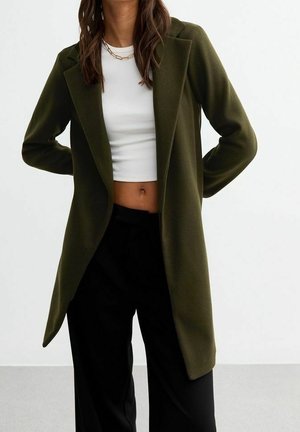 Woman wearing an olive green coat over a white crop top and black pants, standing with hands behind her back against a plain background.