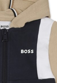 Haut à capuche zippé avec des panneaux marine et beige, tissu texturé et lettrage blanc en logo. Comprend une capuche et des rayures contrastantes.