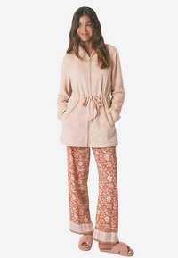 Fleece jas met ritssluiting in zachtroze, met een taillekoord; gecombineerd met bruine broek met bloemenprint en beige, pluizige pantoffels.