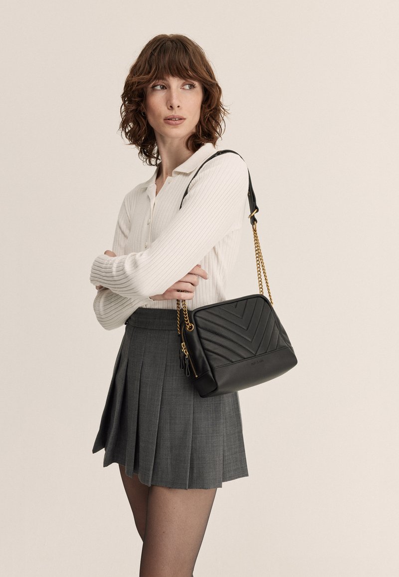 Bolso de cuero negro con acolchado en forma de chevron, correa de cadena dorada y compartimentos con cremallera. Se lleva con una camiseta blanca de canalé y una falda plisada gris.