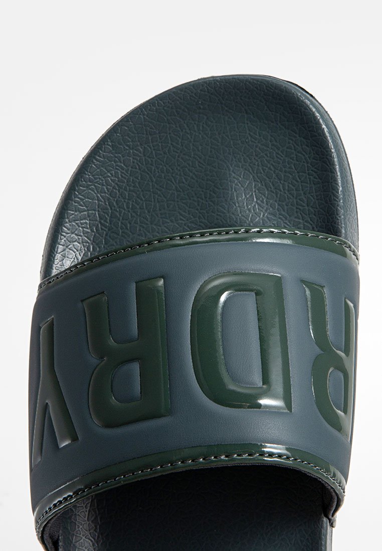 Gros plan sur une sandale en plastique vert foncé avec une semelle texturée et des lettres en relief écrivant "BURRY" sur la bride.