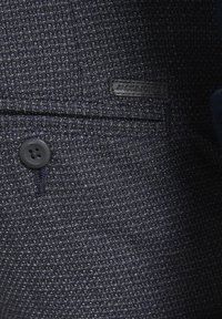 Mørkeblå tekstureret stof med et vævet mønster; har en rund sort knap og et sort lædermærke med "JACK & JONES" præget.