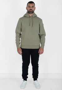 Felpa in cotone verde oliva con coulisse regolabili, polsini e orlo a coste, abbinata a pantaloni jogger neri e sneakers bianche.