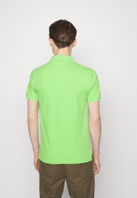 Polo shirt in cotone verde con maniche corte e colletto, caratterizzato da un colore uniforme e un orlo dritto. Dettagli visibili sullo scollo posteriore.