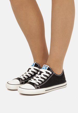 Gambe che indossano scarpe da ginnastica basse in tela nera con punta in gomma bianca, lacci bianchi e etichette con logo blu su sfondo bianco.
