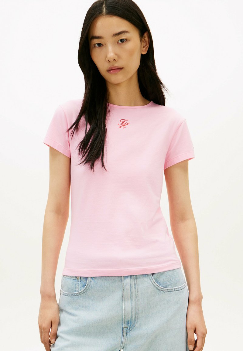 Femme aux longs cheveux foncés portant un t-shirt rose avec des initiales rouges et un jean en denim bleu clair, debout devant un fond blanc uni.