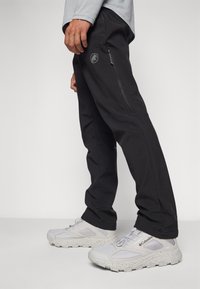 Schwarze Herrenhose mit einer Reißverschlusstasche, kombiniert mit hellgrauen Sneakern. Die Sneaker haben eine strukturierte Sohle und ein Logo an der Seite.
