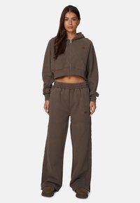 Haut à capuche court marron avec fermeture éclair et logo, associé à un pantalon de survêtement marron ample doté d'une taille élastique et de poches latérales.