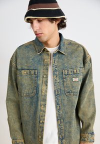 Camicia di jeans con una tonalità sbiadita di blu e giallo, dotata di due tasche frontali e chiusura con bottoni. Indossata sopra una maglietta bianca; persona con un cappello.
