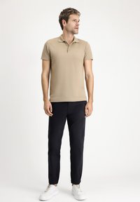 Beżowa koszulka polo z krótkimi rękawami, z zapięciem na trzy guziki, o teksturowanej tkaninie; zestawiona z ciemnymi spodniami i białymi trampkami.