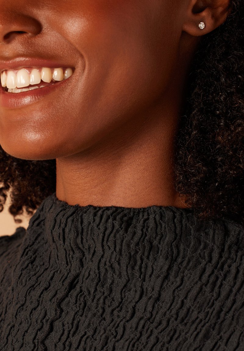 Pull noir texturé avec un col haut et festonné. Le mannequin affiche un sourire chaleureux, agrémenté de petites boucles d'oreilles en forme de clou rond.