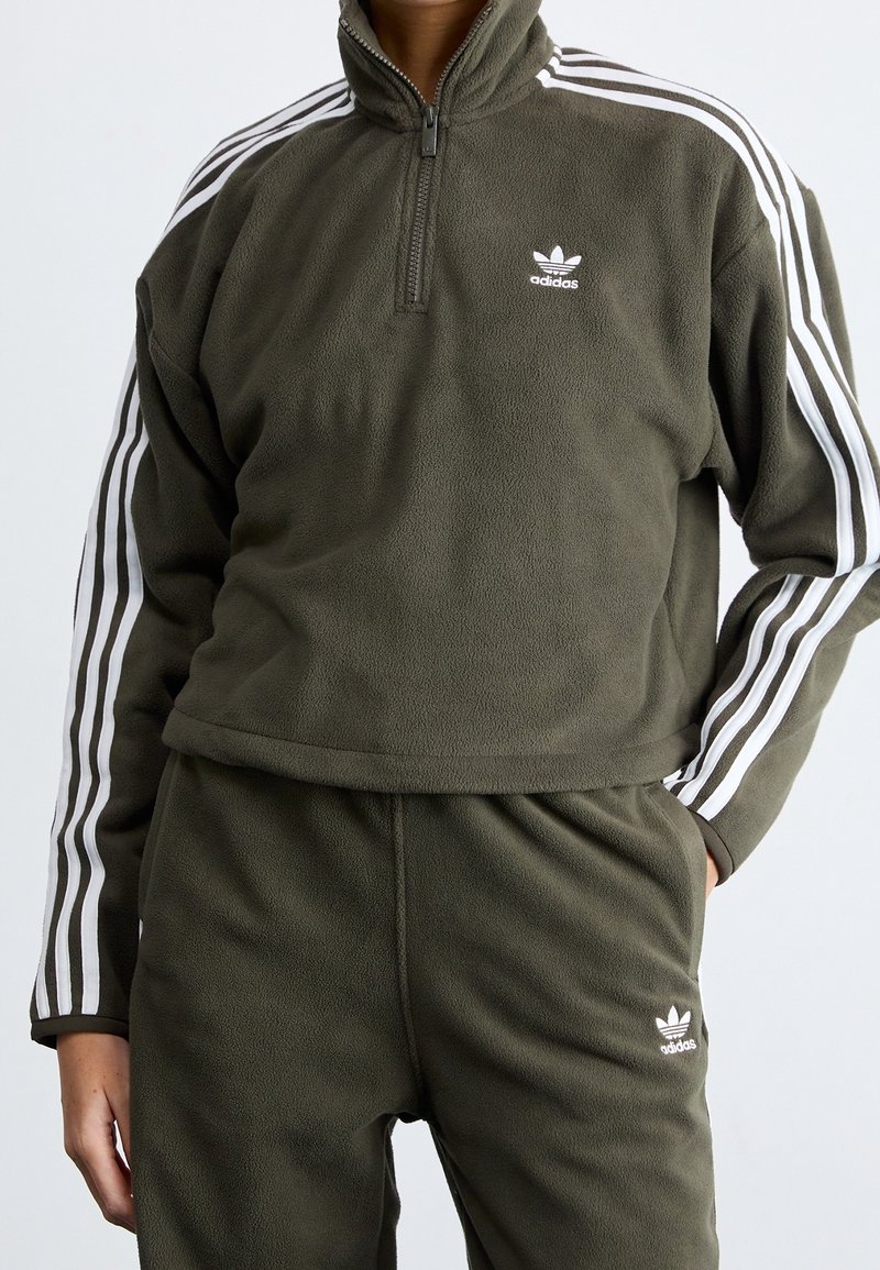 Fleecepullover i olivgrön med dragkedja i halsen, prydd med vita Adidas-ränder på ärmarna och logotyp på bröstet. Mjuk textur, avslappnad passform.