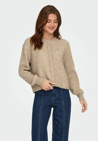 Beżowy sweter z dzianiny z okrągłym dekoltem, przednimi guzikami, prążkowaną teksturą i opadającymi ramionami, w połączeniu z ciemnoniebieskimi spodniami typu straight-leg.