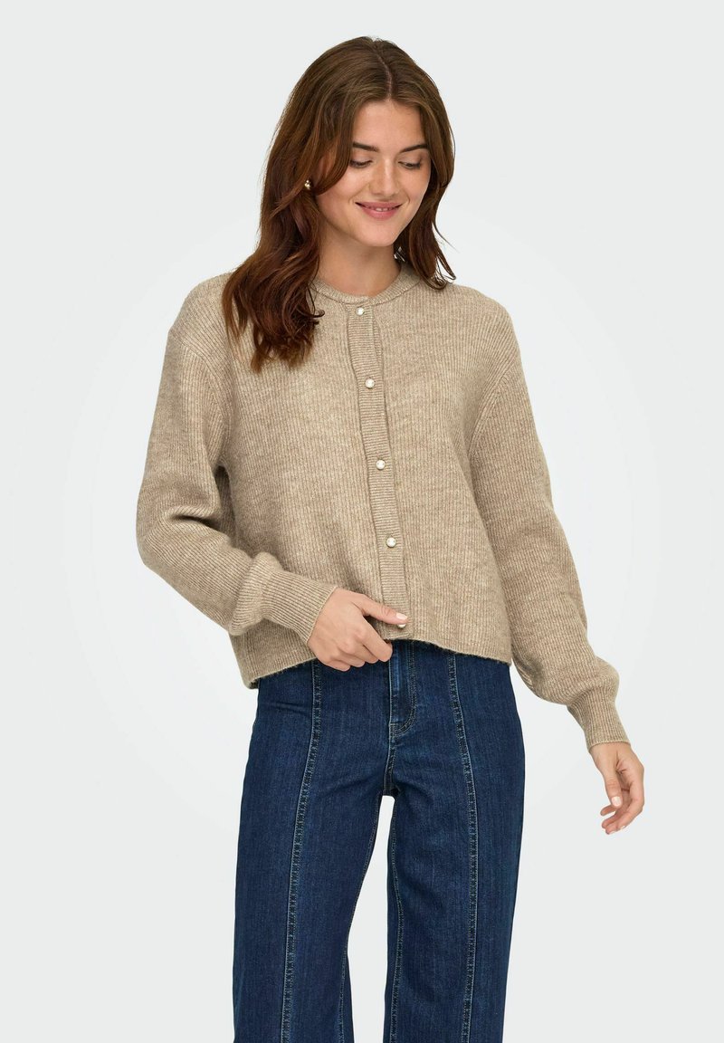 Beżowy sweter z dzianiny z okrągłym dekoltem, przednimi guzikami, prążkowaną teksturą i opadającymi ramionami, w połączeniu z ciemnoniebieskimi spodniami typu straight-leg.