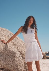 Haut blanc sans manches en tissu texturé et jupe blanche superposée avec une broderie complexe, sur fond de ciel bleu clair.