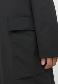 Čierna bunda so štruktúrovaným dizajnom, ktorá má na boku veľkú plochú kapsu. Hladká textura s matným povrchom.