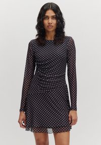 Zwart jurkje met polka dots, lange mouwen, doorzichtige stof, geplooide zijkant en een vloeiende zoom. Het patroon bestaat uit witte stippen.