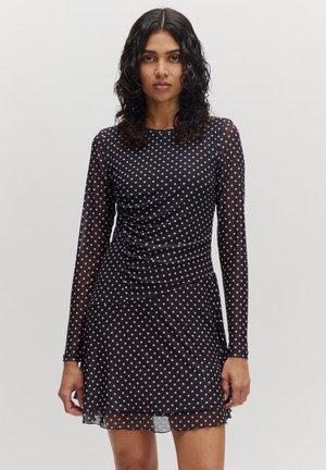 Zwart jurkje met polka dots, lange mouwen, doorzichtige stof, geplooide zijkant en een vloeiende zoom. Het patroon bestaat uit witte stippen.