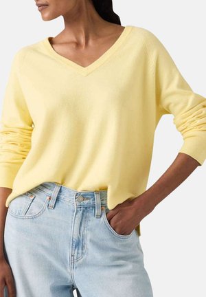 Femme portant un pull à col en V jaune pâle avec les manches retroussées, légèrement rentrée dans un jean taille haute bleu clair, la main dans la poche.