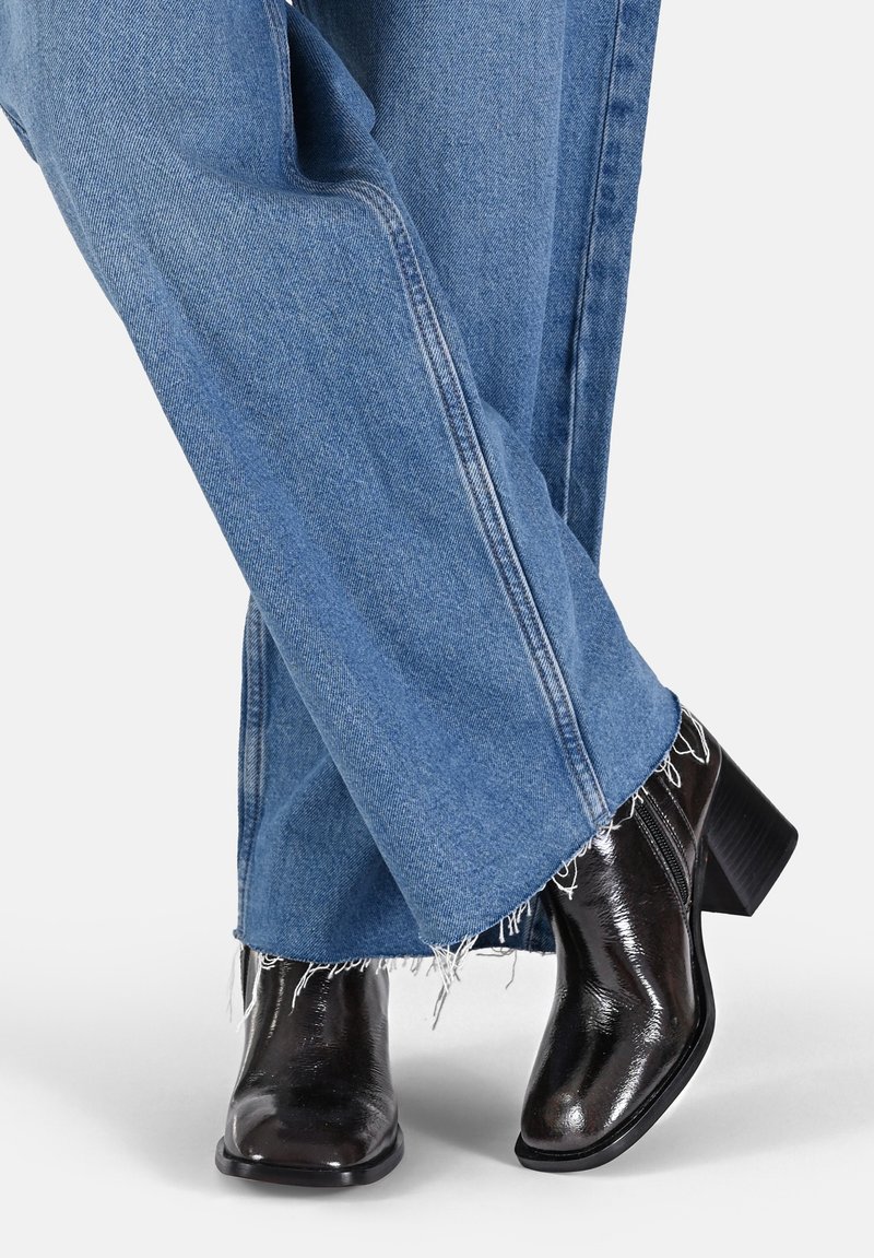 Bottines noires à talons avec une finition brillante, associées à un jean taille haute bleu clair à jambes larges, avec des ourlets effilochés et des détails de surpiqûres sur les côtés.