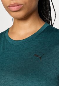 Teal atletisk t-skjorte med rund hals, med en subtil svart Puma-logo på brystet. Materialet ser jevnt ut med en liten tekstur.