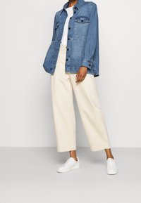 Chaqueta de mezclilla azul clara con cierre de botones y dos bolsillos en el pecho, combinada con pantalones de pierna ancha color crema y zapatillas blancas.