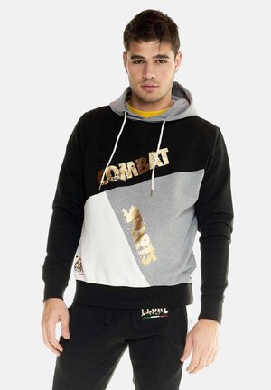 Jonge man draagt een zwarte, grijze en witte hoodie met gouden "COMBAT SPORTS" tekst, gecombineerd met zwarte trainingsbroek tegen een effen achtergrond.