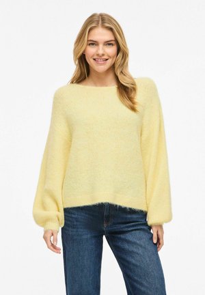 Femme souriante, portant un pull ample jaune clair à manches bouffantes et un jean bleu foncé, debout devant un fond blanc.