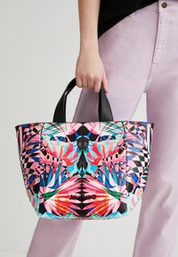 Sac fourre-tout coloré en matériau synthétique, présentant un motif floral vibrant, des accents noirs et des poignées courtes. Forme carrée.