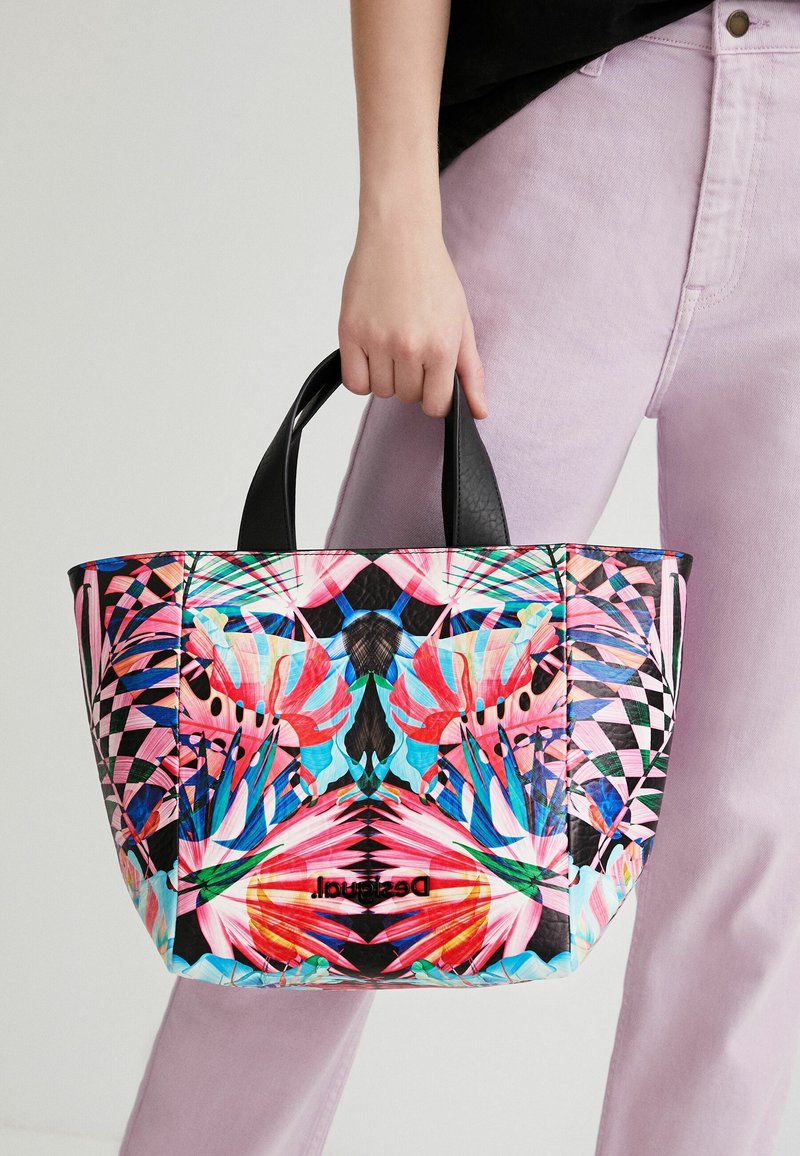 Sac fourre-tout coloré en matériau synthétique, présentant un motif floral vibrant, des accents noirs et des poignées courtes. Forme carrée.