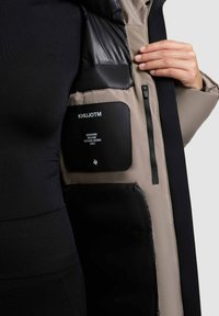 Puffer jacket interior showing a beige lining, black accents, a zippered pocket, and a black label with brand and establishment details embossed.