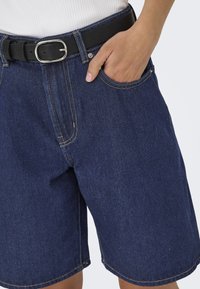 Shorts en denim bleu foncé, à coupe droite, avec des coutures contrastées et une ceinture noire avec une boucle en argent, ainsi que des poches latérales.