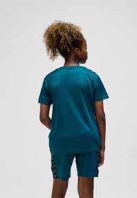 Turkos atletisk tröja med avslappnad passform och korta ärmar, kombinerad med matchande shorts. Har svarta chevronmönster på sidan. Mjuk textur.