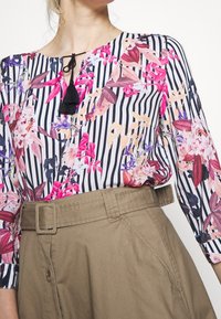 Bluzka w kwiatowy wzór z pionowymi czarnymi i białymi paskami, z okrągłym dekoltem i czarnym frędzlem. Połączona z khaki spódnicą.