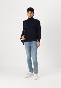 Marineblauer Rollkragenpullover, hellblaue Jeans, weiße Sneakers; Modell hält eine schwarze Umhängetasche und steht vor einem schlichten weißen Hintergrund.