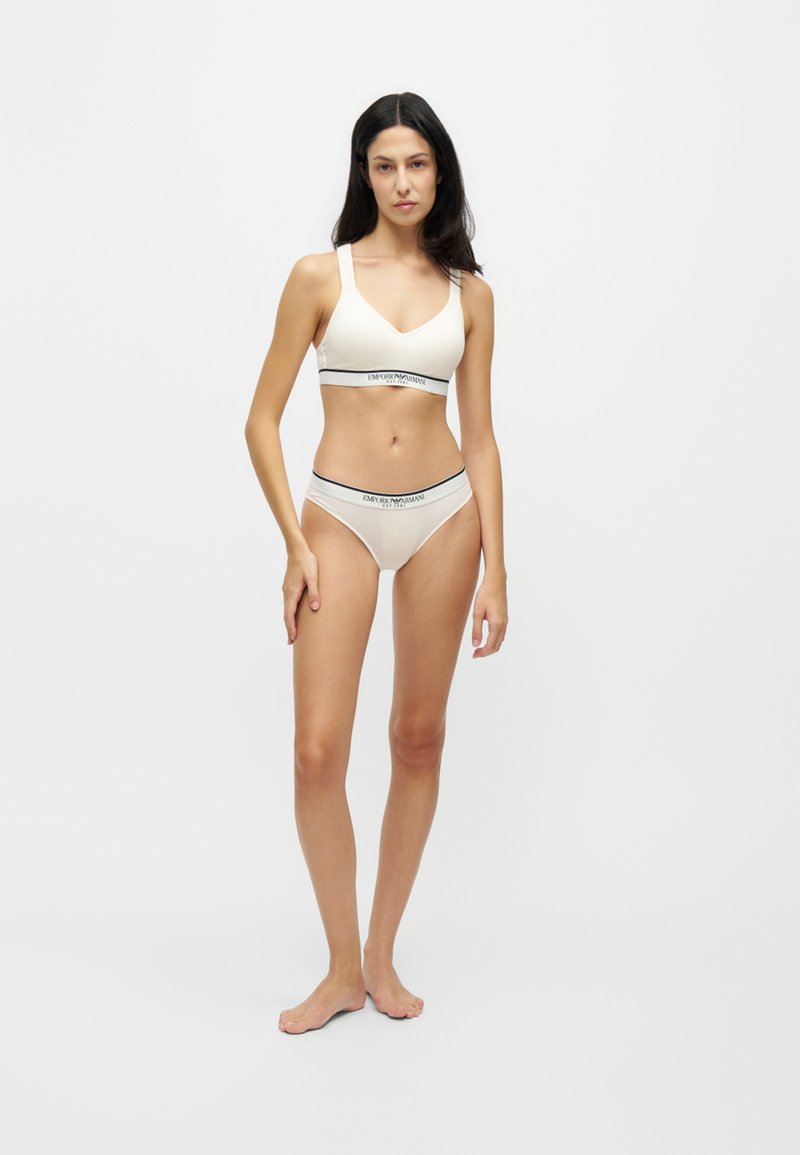 Femme debout pieds nus portant un soutien-gorge blanc de marque et un slip assorti sur un fond clair uni.