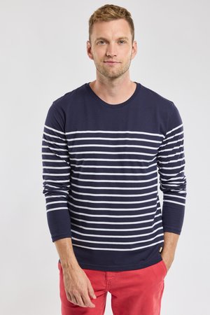 Uomo con capelli biondi corti che indossa una maglia a maniche lunghe blu navy con strisce orizzontali bianche e pantaloni rossi, in piedi contro uno sfondo chiaro e uniforme.