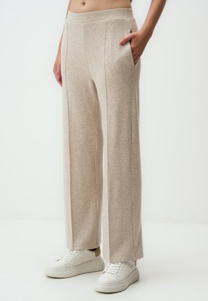 Beige weitgeschnittene Hose aus weichem Stoff mit glatter Textur, ausgestattet mit Fronttaschen und hoher Taille, kombiniert mit weißen Sneakers.