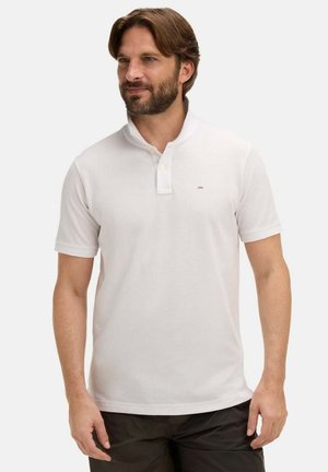 Homme portant un polo blanc à manches courtes avec col, debout devant un fond uni, regardant légèrement sur le côté.